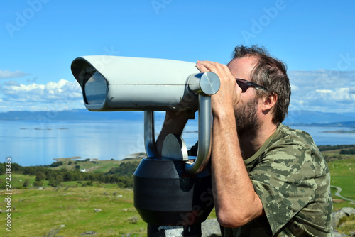 A man looks at the panoramic binoculars from the top of the hill. View of the sea. Blue sky. Sunny day.