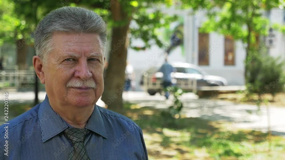 Brave senior man with sharp eyes standing in a green street in summer in slo-mo Brave senior man with sharp eyes standing in a green street in summer in slo-mo
