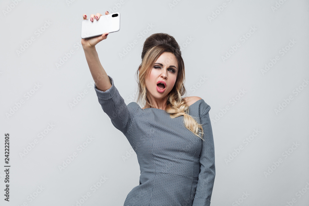 Obraz premium Selfie time! Happy beautiful blogger young girl wearing in grey dress with pigtail on head standing, winking with sexy open mouth and making selfie. Indoor, isolated, studio shot, grey background