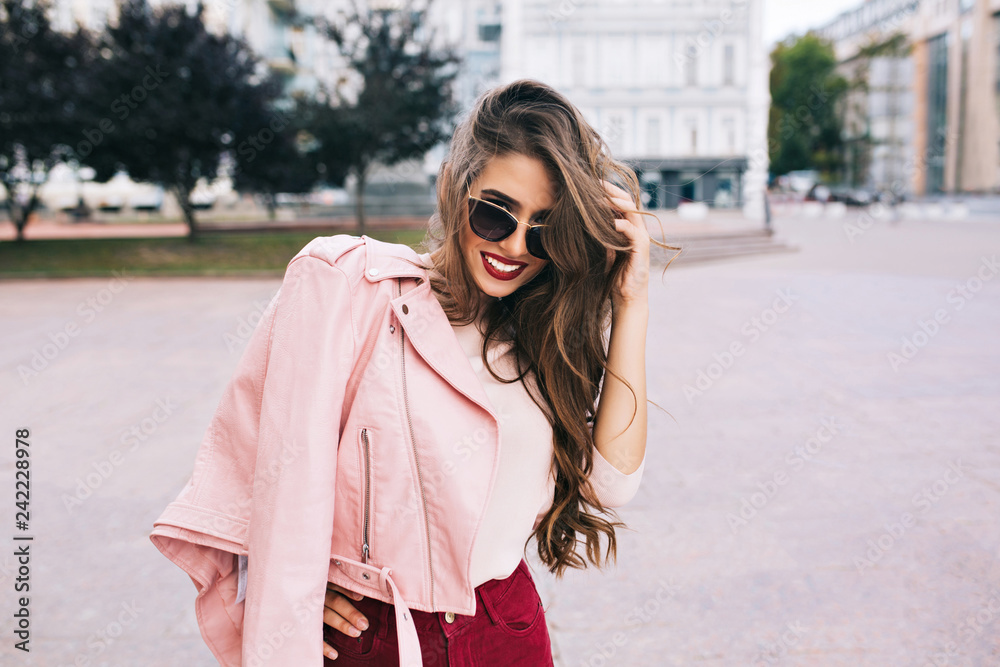 Attractive girl in sunglasses with snow-white smile is posing to camera in city. She has pink jacket on shoulders, long hairstyle.