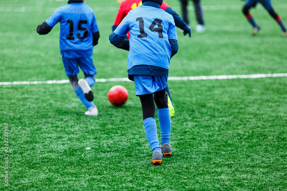 Boys in red and blue sportswear plays soccer on green grass field. Youth football game. Children sport competition, kids plays outdoor, winter activities, training Boys in red and blue sportswear plays soccer on green grass field. Youth football game. Children sport competition, kids plays outdoor, winter activities, training