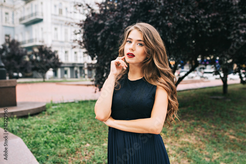 Portrait of serious elegante girl in the city park. She has long hair, black dress, vinous lips and looks wistfully.