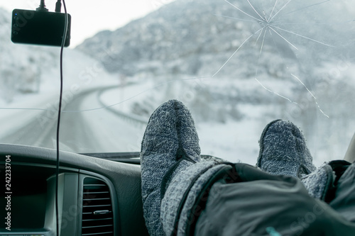 human feet in warm boots on the panel of the car, on the background of broken windshield and winter slippery snow-covered road in the highlands