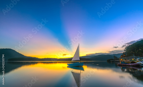 Lonely sailboat in the lake during beautiful sunset with sun rays clouds shines from sky colorful reflecting off the mystical lake to end the day