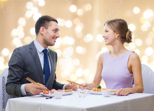 people and food concept - smiling couple eating sushi rolls by chopsticks at restaurant over festive lights on beige background