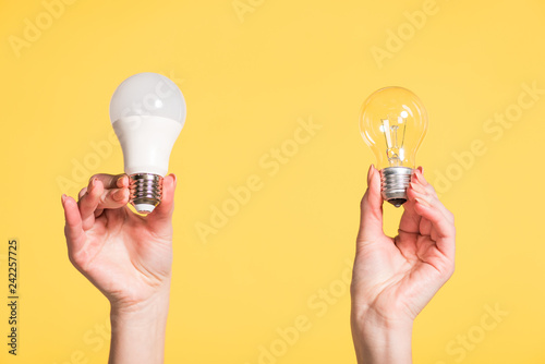 cropped view of female hands choosing between led and fluorescent lamps isolated on yellow, energy efficiency concept