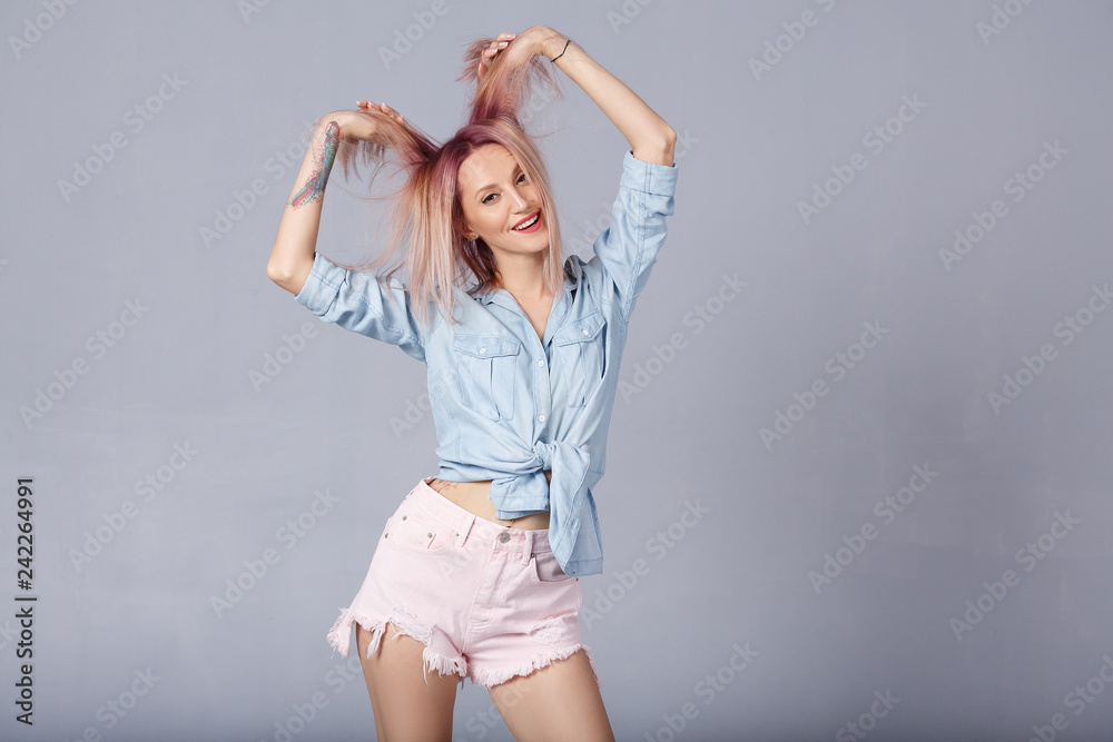 Beautiful young woman expresses happy emotions, has broad pleasant smile, glad to new unusual pink color of his hair. Smiling overjoyed female in blue denim shirt enjoys over gray background Beautiful young woman expresses happy emotions, has broad pleasant smile, glad to new unusual pink color of his hair. Smiling overjoyed female in blue denim shirt enjoys over gray background