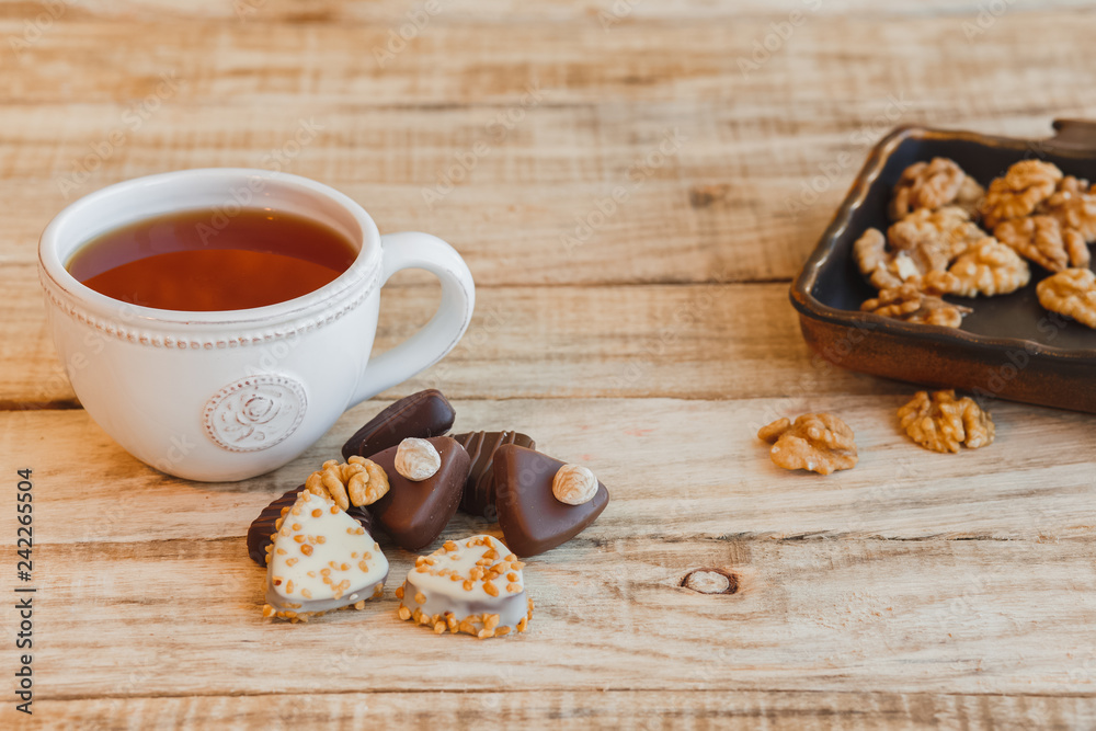 Cup of hot black tea with chocolate candy and walnuts on wooden table, selective focus.