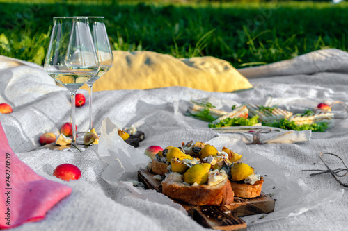 Picnic in forest on sunny meadow, blanket, wicker basket, wine glasses, bruschetta with cheese and pear, snacks, fruit, apples, bread. Concept of romantic lunch date in picturesque forest
