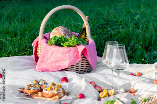 Picnic in forest on sunny meadow, blanket, wicker basket, wine glasses, bruschetta with cheese and pear, snacks, fruit, apples, bread. Concept of romantic lunch date in picturesque forest