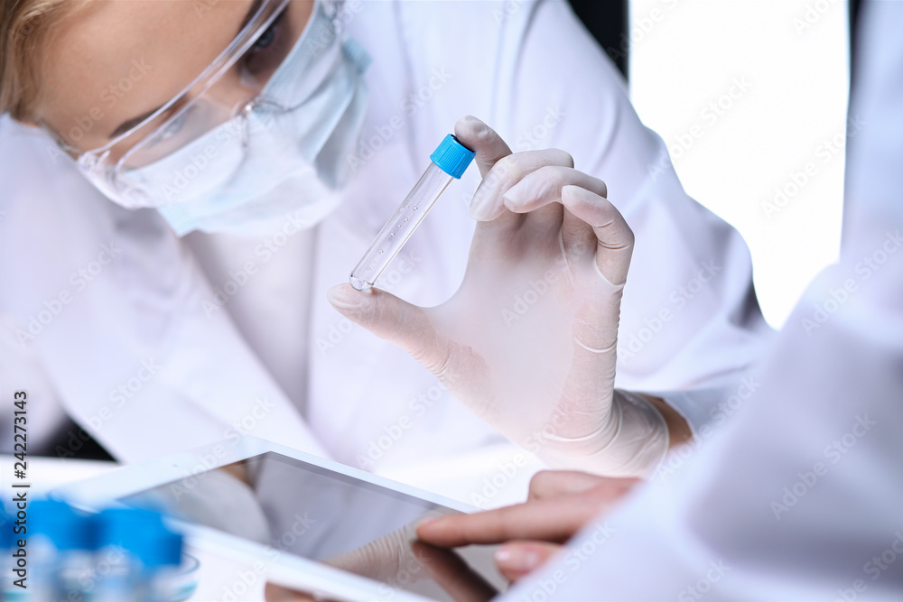 Closeup of scientific research team with clear solution in laboratory. Blonde female chemist holds test tube of glass while her colleague checks results with tablet pc. Blood test, medicine or Closeup of scientific research team with clear solution in laboratory. Blonde female chemist holds test tube of glass while her colleague checks results with tablet pc. Blood test, medicine or