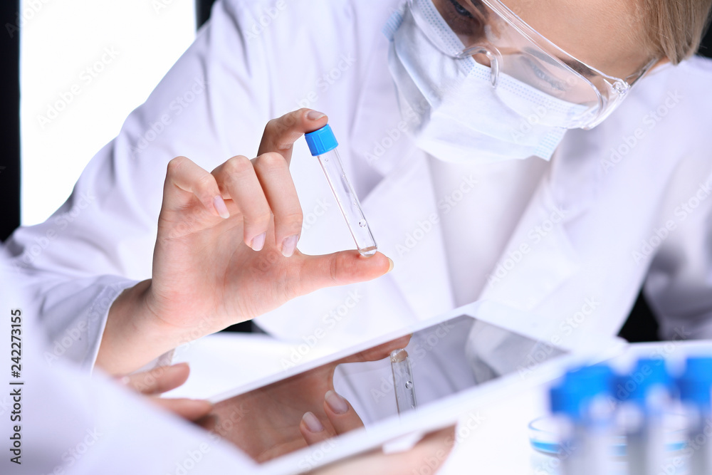 Closeup of scientific research team with clear solution in laboratory. Blonde female chemist holds test tube of glass while her colleague checks results with tablet pc. Blood test, medicine or Closeup of scientific research team with clear solution in laboratory. Blonde female chemist holds test tube of glass while her colleague checks results with tablet pc. Blood test, medicine or