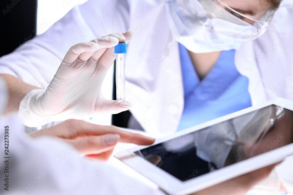 Closeup of scientific research team with clear solution in laboratory. Blonde female chemist holds test tube of glass while her colleague checks results with tablet pc. Blood test, medicine or Closeup of scientific research team with clear solution in laboratory. Blonde female chemist holds test tube of glass while her colleague checks results with tablet pc. Blood test, medicine or