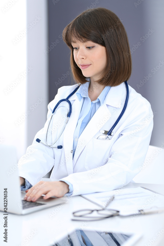 Doctor typing on laptop computer while sitting at the white table in hospital office. Physician at work. Medicine and healthcare concept Doctor typing on laptop computer while sitting at the white table in hospital office. Physician at work. Medicine and healthcare concept