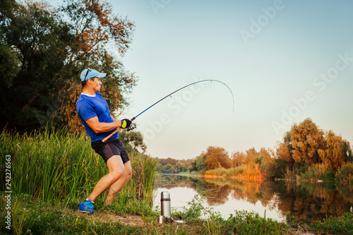 Young man fishing on river at sunset. Happy excited fiserman pulling rod with fish.