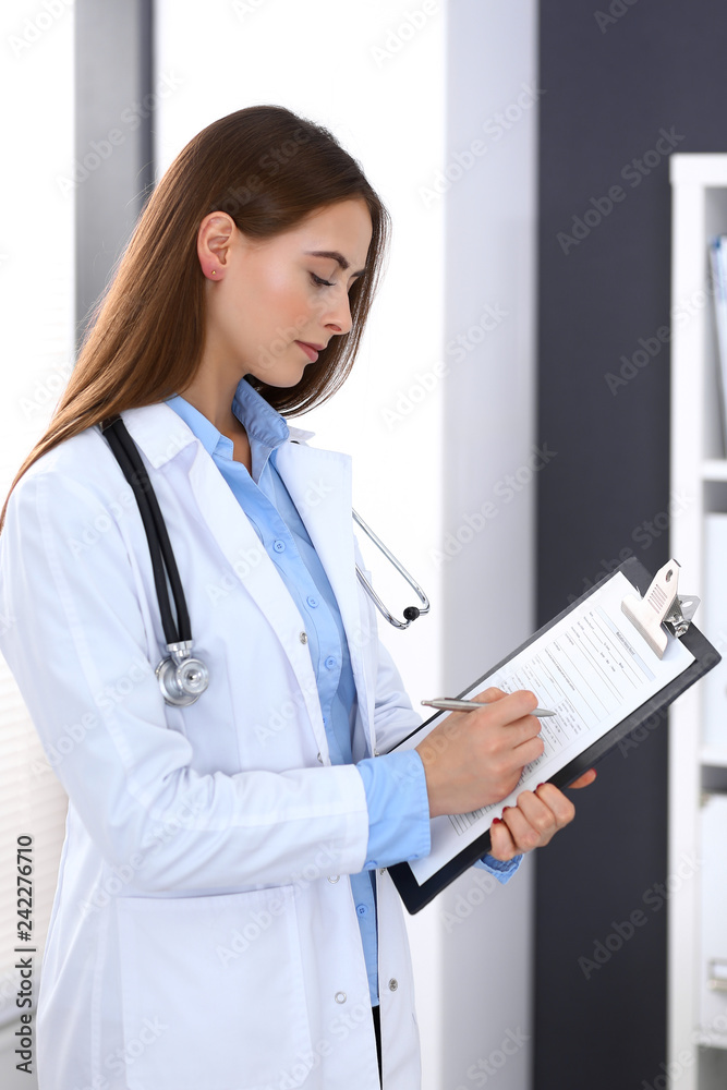 Doctor woman filling up medical form while standing near window in hospital office. Happy physician at work. Medicine and health care concept Doctor woman filling up medical form while standing near window in hospital office. Happy physician at work. Medicine and health care concept