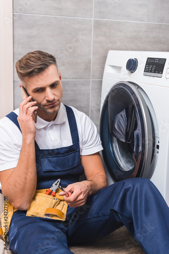 adult repairman holding screwdriver and talking on smartphone while repairing washing machine in bathroom