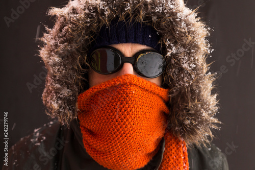 Portrait of a single male winter adventurer wearing a warm green coat with fur hood, a blue ski cap, an orange scarf and black retro style goggles