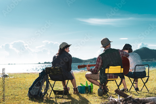 Camping group of friends asian people together, teenagers travel with backpack,tent man playing guitar song relaxing on vacation time holiday sitting on chairs near river and mountain view.