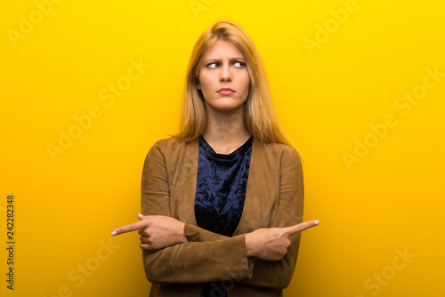 Blonde girl on vibrant yellow background pointing to the laterals having doubts