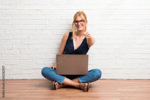 Blonde girl sitting on the floor with her laptop giving a thumbs up gesture and smiling on white brick wall background