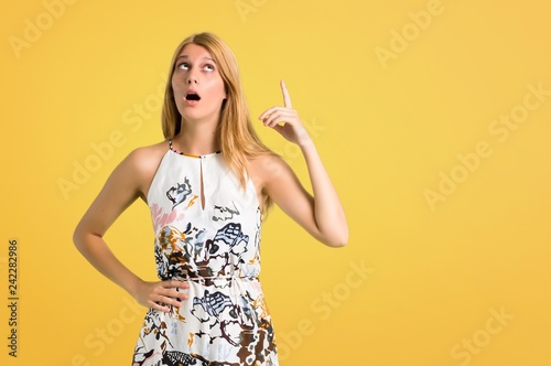 Blonde young girl in a summer dress standing and thinking an idea pointing the finger up on yellow background