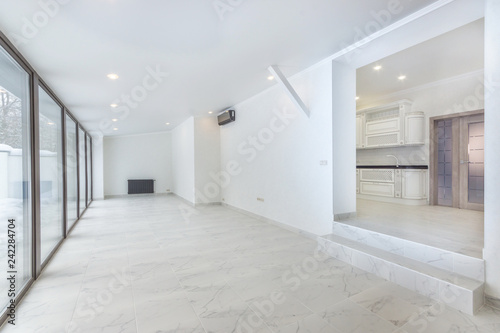 Empty white room with tiled floor and floor to ceiling window. Sliding doors to the back yard. Large doorway to the kitchen