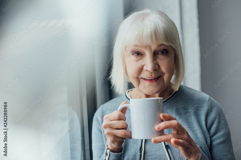 smiling senior woman holding cup of coffee at home and looking at camera smiling senior woman holding cup of coffee at home and looking at camera