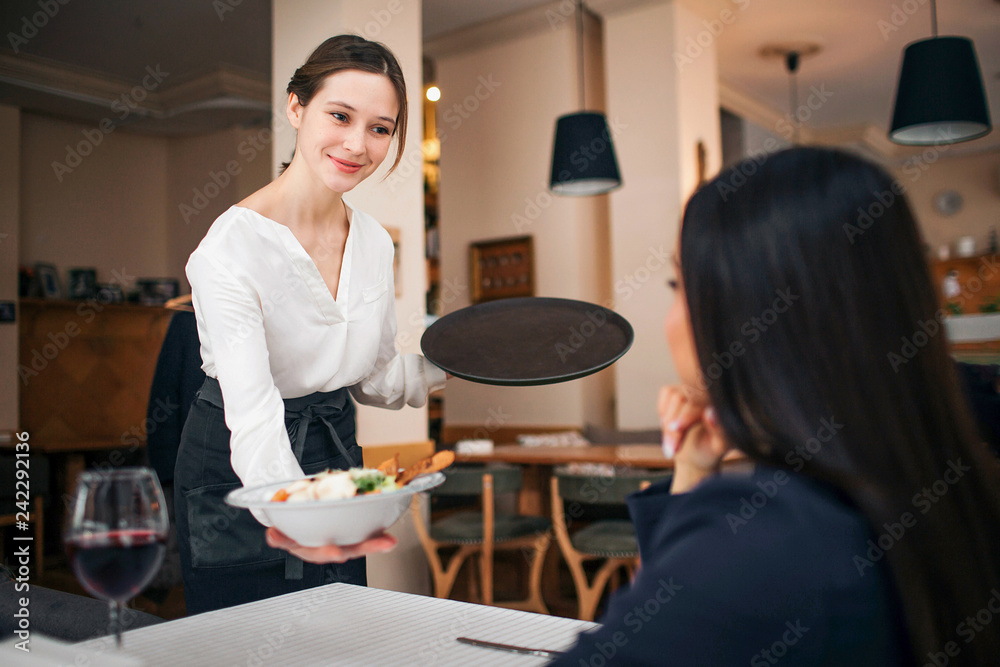 Pleasant young waitress stand at table with sitting customer and show her bowl of salad. She smiles. Female customer look at it. They are in restaurant. Pleasant young waitress stand at table with sitting customer and show her bowl of salad. She smiles. Female customer look at it. They are in restaurant.