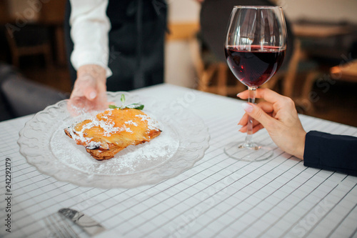 Cut view of woman's hand holding plate with cake. Customer's hand has glass of red wine. People are inside in restaurant.