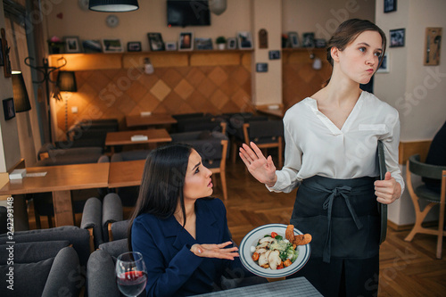 Humble young businesswoman sit at table and show salad bowl to waitress. She refuse to look at it. Young woman in white blouse show stop sign. photo