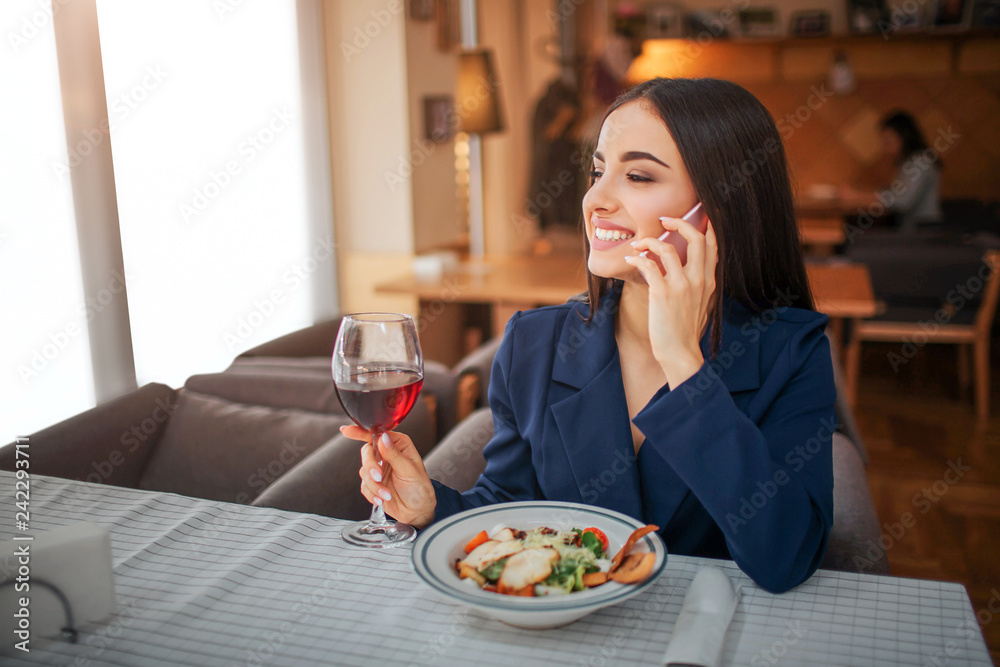 Cheerful young woman sit at table and talk on phone. She smiles. Model hold glass of red wine. Salad bowl stand in front of her at table.