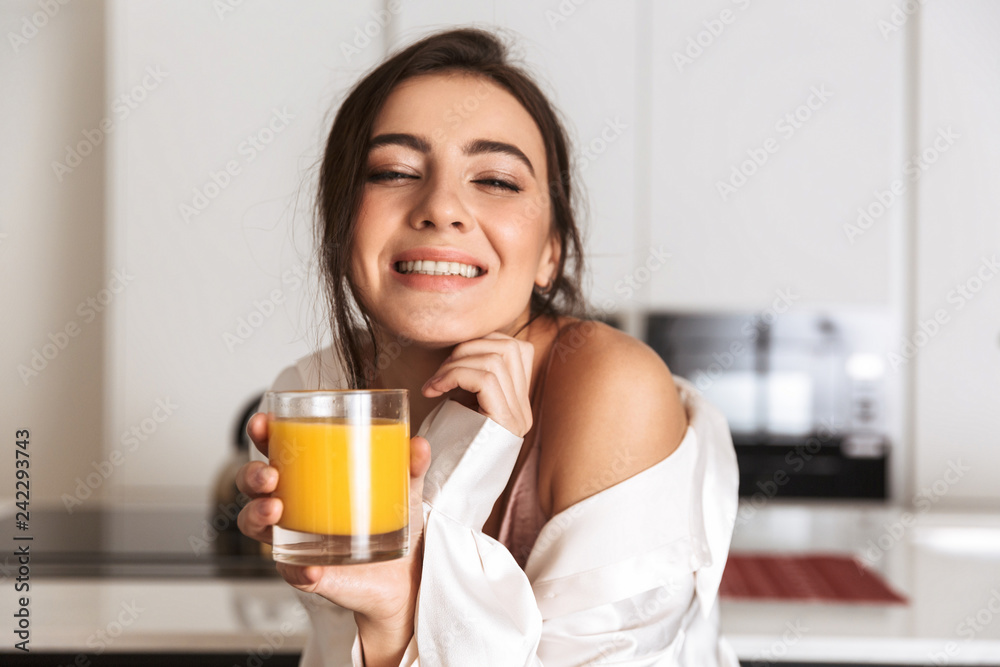 Photo of beautiful woman 30s wearing silk clothing smiling, and drinking orange juice in kitchen Photo of beautiful woman 30s wearing silk clothing smiling, and drinking orange juice in kitchen