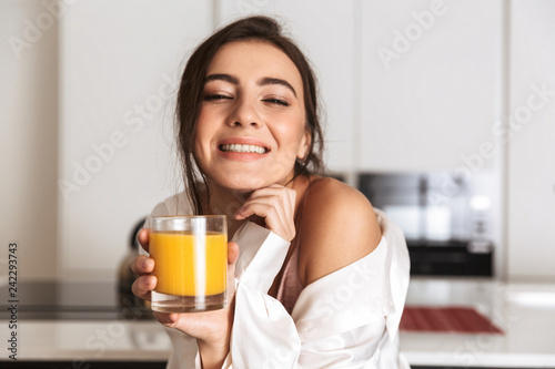 Photo of beautiful woman 30s wearing silk clothing smiling, and drinking orange juice in kitchen