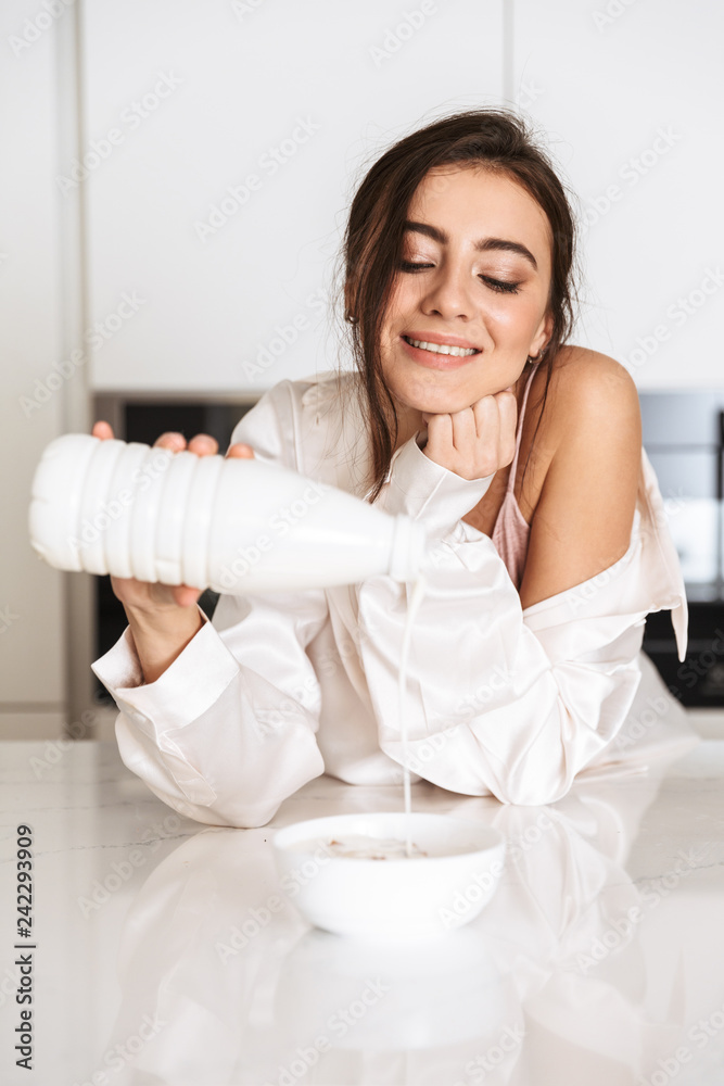 Photo of brunette woman 30s wearing silk clothing eating granola with milk, while having breakfast in apartment Photo of brunette woman 30s wearing silk clothing eating granola with milk, while having breakfast in apartment