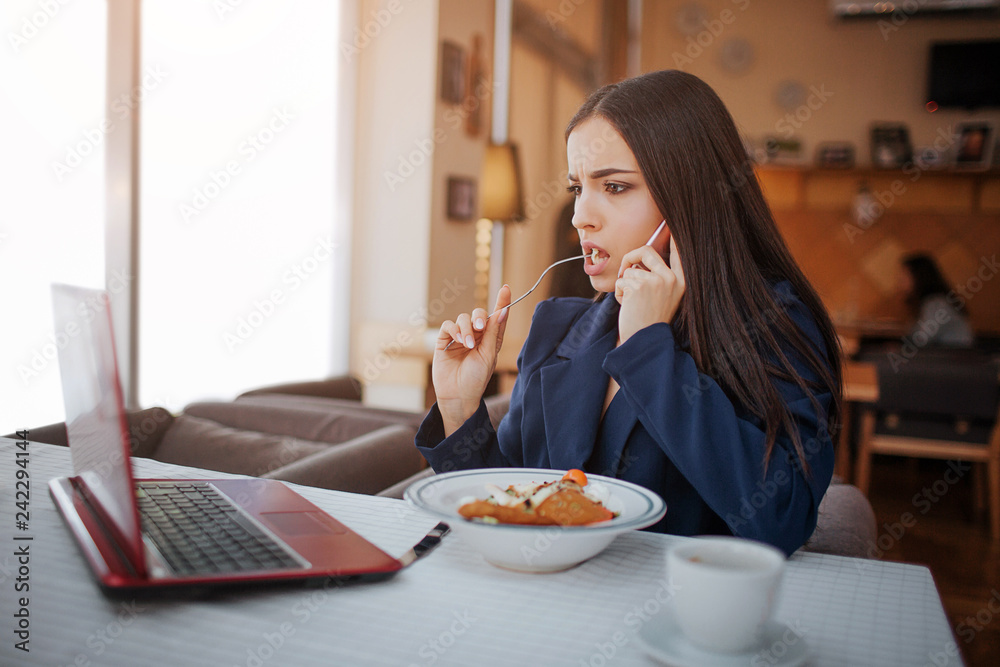 Serious young woman sit at table in restaurant. She talk on phone and look at laptop screen. Model eat salad. Serious young woman sit at table in restaurant. She talk on phone and look at laptop screen. Model eat salad.