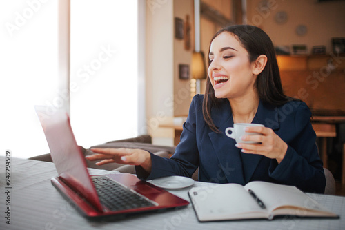 Excited and cheerful young woman sit at table in restaurant. She point on laptop's screen. Model smiling. She has notebook with pen on table.