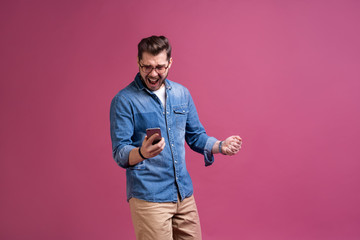 Always in touch. Smiling young man holding smart phone and looking at it. Portrait of a happy man using mobile phone isolated over pink background