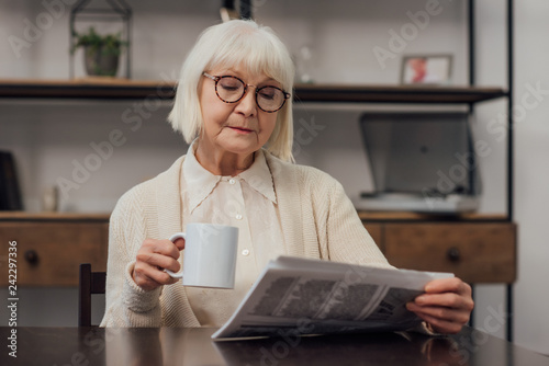 senior woman in glasses sitting at table, drinking coffee and reading newspaper at home