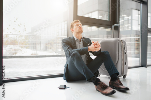 Serious young amn wait for his flight in airport. He lean to window and keep eyes closed. Guy listen to music thrugh headphones. He left phone and suitcase besides himself. photo