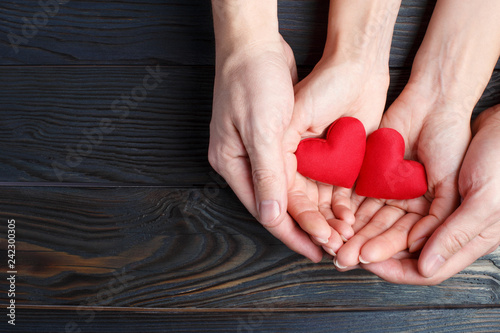 Male and female hands holding two red love hearts on wooden background, view from above