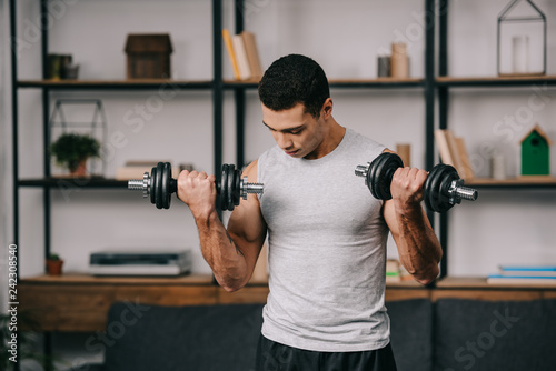 strong mixed race man training with dumbbells in living room