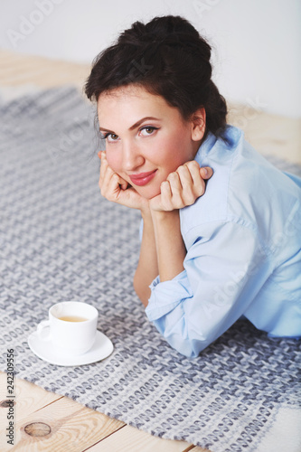 Fotografia Young beautiful brunette woman, wearing blue shirt, lying on a grey woolen blanket with cup of tea