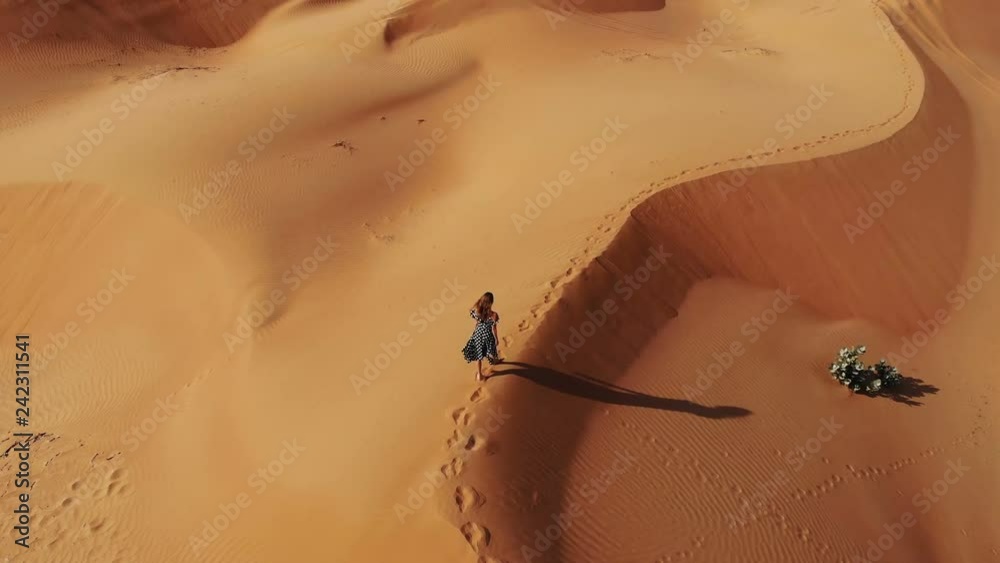 AERIAL. Woman weared in long dress walking in the Dubai desert sand dunes with footsteps in the sand during sunset