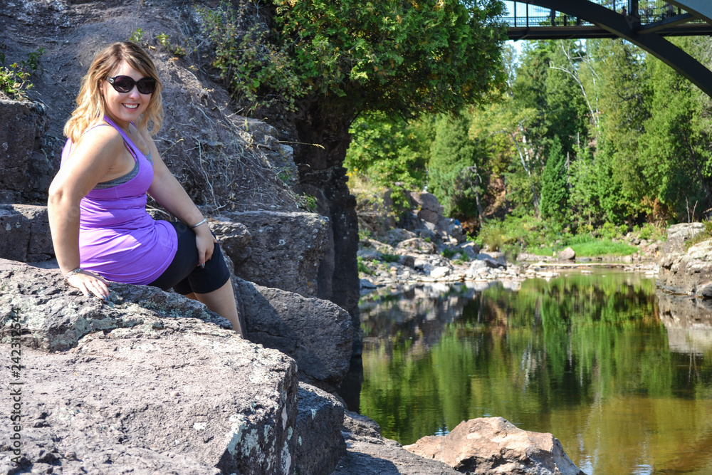 Portrait of a young adult woman wearing sunglasses and smiling sitting on a cliff outdoors near a calm river in Minnesota Portrait of a young adult woman wearing sunglasses and smiling sitting on a cliff outdoors near a calm river in Minnesota