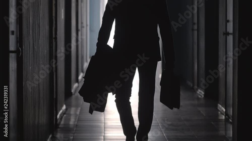 Tilt up shot of silhouette of unrecognizable businessman wearing formal suit and carrying coat and briefcase walking away from the camera along hallway in office center