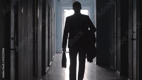Tilt down shot of silhouette of businessman wearing formal suit and carrying coat and briefcase leaving office and walking towards the camera along hallway 