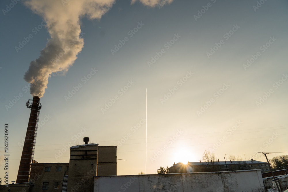 In the setting sun the plane flies against the background of the factory that smokes from the chimney.