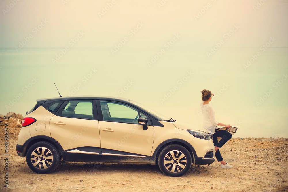 Happy stylish young woman traveler on beach road sitting on white crossover car, holding hat in hand. Banner. Travel, summer vacation, holiday, freedom concept. Digital detox