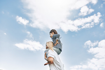 Side view of cute little daughter on a piggy back ride with her happy mother on the sky background. Loving woman and her little girl playing in the park. Mom and kid has fun outside. Mother's day